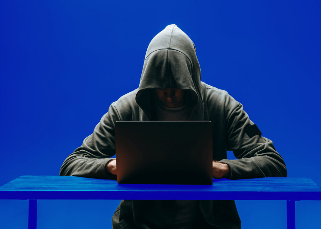 A man wearing a hoodie with cap on sitting at a table looking at his laptop.