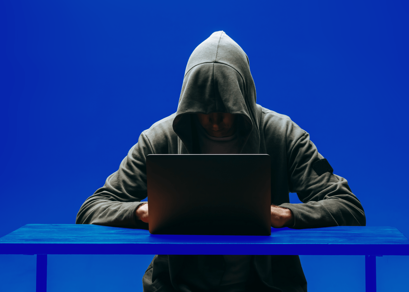 Een man met een hoodie en een pet op zit aan een tafel en kijkt naar zijn laptop.